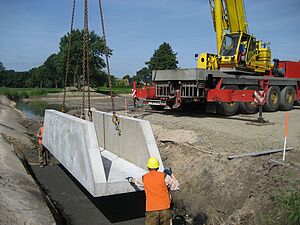 Ein sehr großer Kran auf vierachsigem Tieflader lässt ein Betonfertigteil in eine Baugrube herab. In der Baugrube stehen zwei Bauarbeiter und nehmen das Teil entgegen.