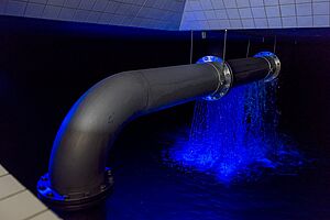 Detail einer Wasserrohrleitung, dahinter ein sprudelnder Wasserschwall, alles in blaues Licht getaucht.