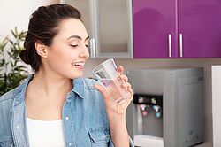 Ein Mensch steht in einem Küchenumfeld und trinkt ein Glas Wasser mit frohem Gesichtsausdruck.