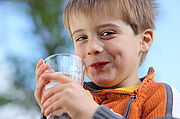 Ein kleiner Junge hält ein Glas Wasser in den Händen und grinst verschmitzt.