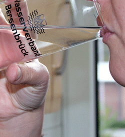 Ein Mensch trinkt Wasser aus einem Glas mit dem Aufdruck "Wasserverband Bersenbrück" (Nahaufnahme, nur Mund, Hand und Glas sind zu sehen).