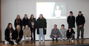 Elf Schüler*innen und ihr Lehrer stehen vor der Projektion ihrer Präsentation.