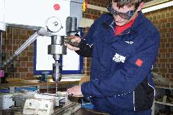 Ein junger Mensch mit blauer Arbeitsmontur und Schutzbrille bedient einen Bohrständer.