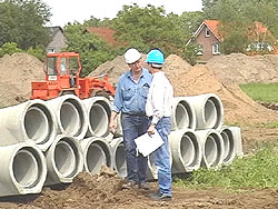 Zwei Arbeiter stehen vor einem Stapel Kanal-Rohre; im Hintergrund aufgeschüttete Sandhügel und ein Radlader.