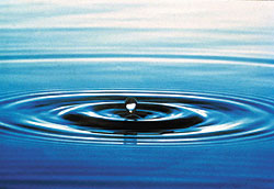 Ein Tropfen fällt auf eine blau spiegelnde Wasseroberfläche und erzeugt ringförmige Wellen