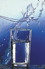 Ein zu zwei Dritteln gefülltes Wasserglas vor blauem Hintergrund, von hinten spritzt ein Schwall Wasser über das Glas.